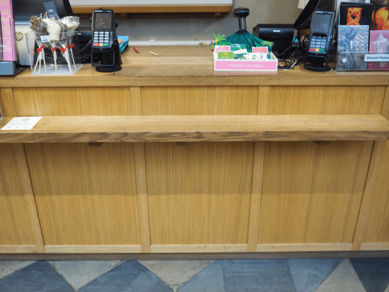 The shop counter with the till on the far left, and the card reader next to the till. There are postcards and sweets along the tip of the counter. There is also a lower lip at the front of the counter.