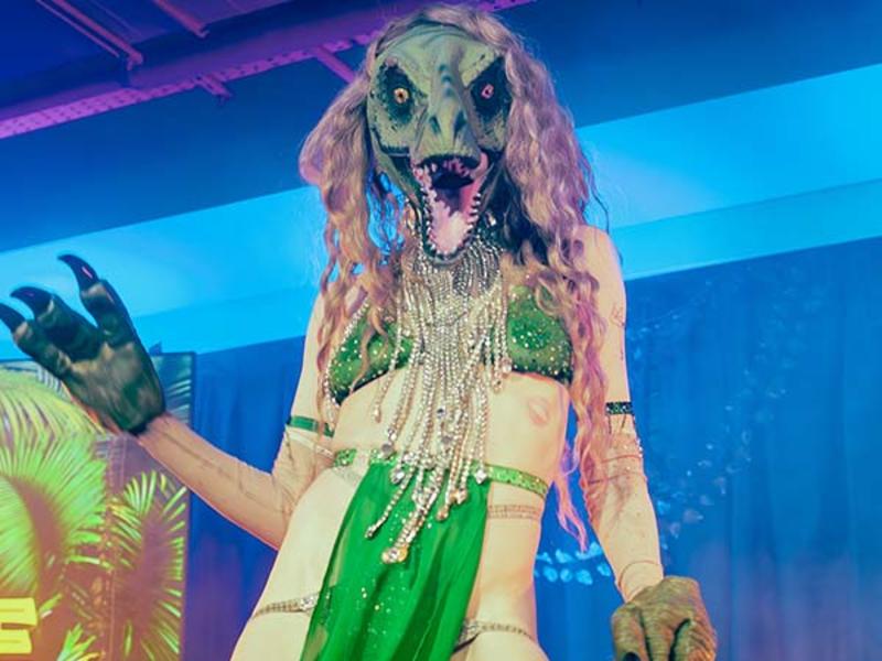 A performer in a detailed dinosaur mask and claws, wearing a glittery green and silver costume, poses on stage under colorful lighting.