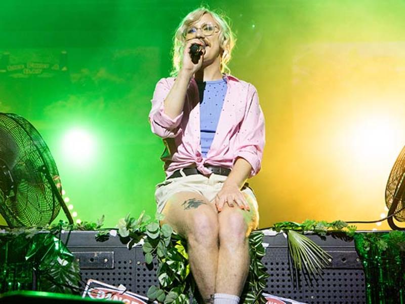 A performer with blonde hair, glasses, and a microphone sits on the edge of the stage, wearing a pink shirt tied at the waist, a blue undershirt, and beige shorts, with stage lights glowing green and yellow behind them.