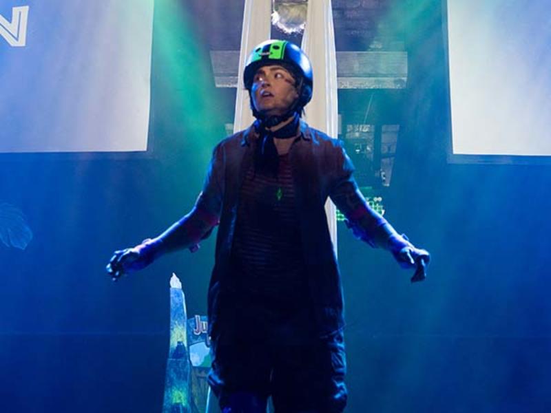 A performer in dark clothing and a helmet looks upward on stage, dramatically lit with blue and green spotlights.