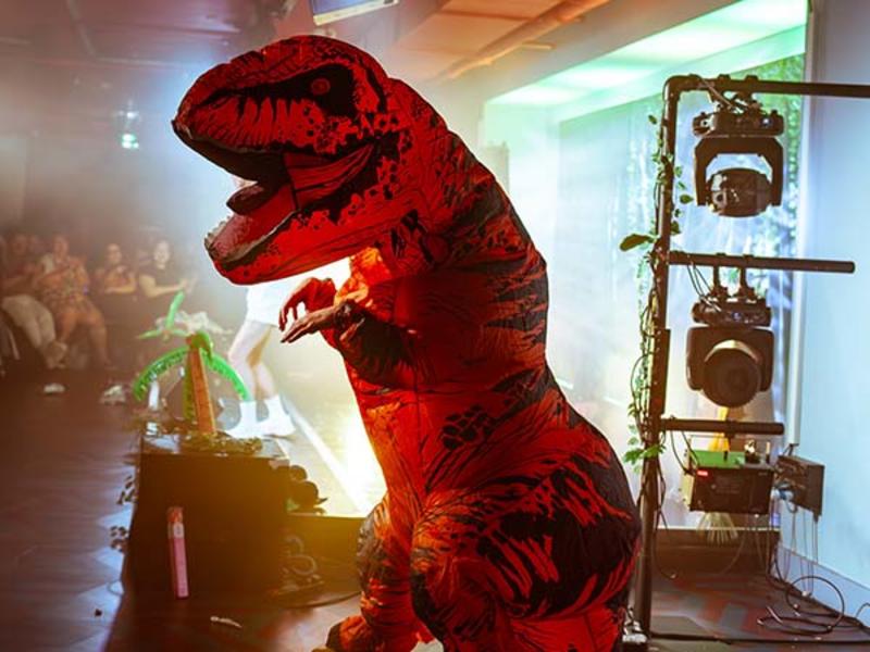 A performer wearing a humorous dinosaur costume performs on stage against a bright light.